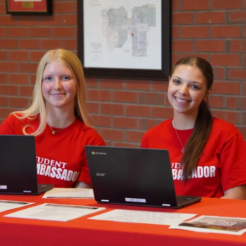 Two female students sitting at a table smiling with chromebooks in front of them.